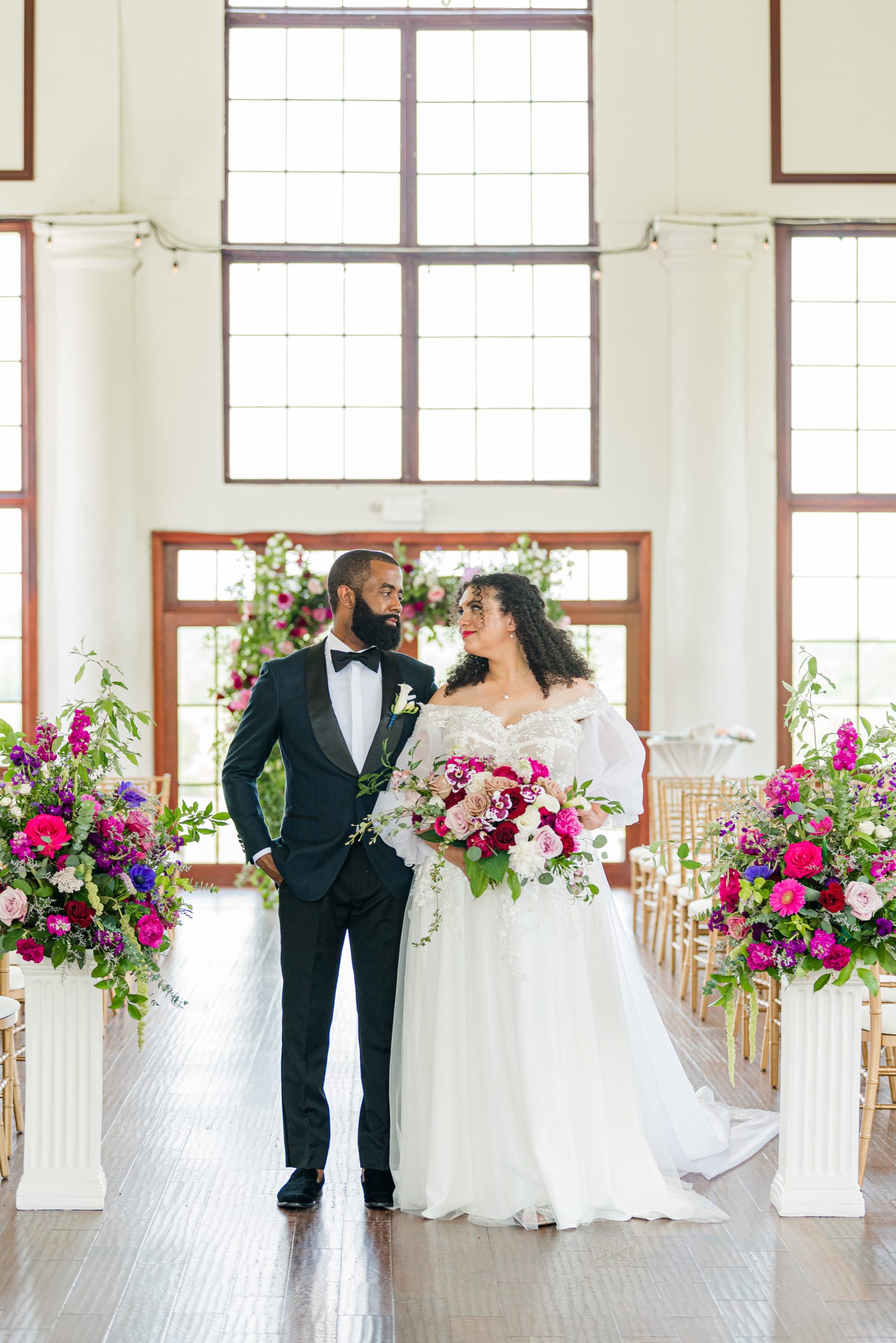 Couple dressed in wedding attire at their ceremony cite covered in bright colored pink and red flowers.